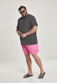 T-shirt nera con motivo grafico bianco, pantaloni corti rosa e sandali neri. Modello in posa con le braccia leggermente piegate, indossando occhiali da sole scuri.