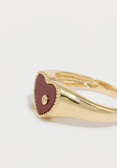 Zlatý prsteň so srdcovým červeným smaltovaným vložením, zdobený textúrovaným okrajom a malým priehľadným drahokamom uprostred.