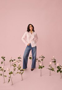 Frau trägt ein rosa gestreiftes Hemd und blaue Jeans, steht zwischen Glasvasen mit rosa Rosen vor einem hellrosa Hintergrund.