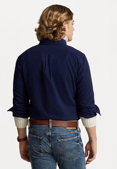 Camisa azul-marinho com mangas dobradas, feita de tecido suave. Cinto de couro castanho emparelhado com jeans azuis desbotados, mostrando um corte clássico.