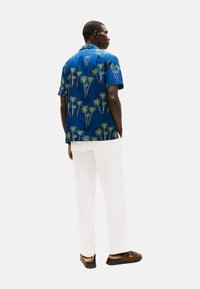 Camisa de manga corta azul con estampado de palmeras, combinada con pantalones blancos y sandalias marrones. La camisa cuenta con un cuello tipo polo y un corte relajado.
