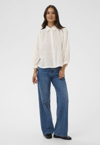 Chemise blanche à boutons avec une subtile texture en zigzag, associée à un jean en denim bleu à jambes larges et des chaussures noires, sur un fond uni.