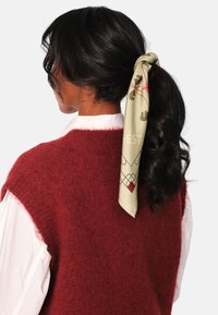 Rød, fluffy, ermeløs genser kombinert med en hvit skjorte med krage. Et grønt mønstret skjerf er knyttet i håret, med blomster- og geometriske design.