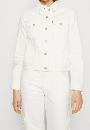 Kvinde iført hvid denimjakke med knapper og matchende hvide cargobukser, stående foran en ensfarvet baggrund.