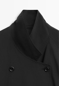 Zwarte double-breasted jas met brede reverskraag en twee zichtbare glanzende zwarte knopen aan de voorkant.