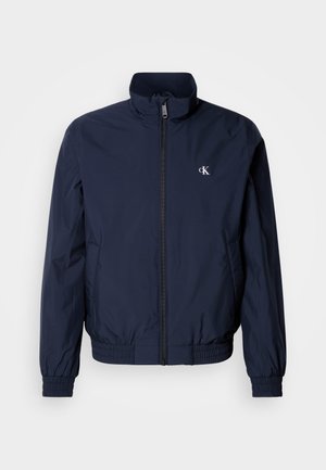 Navy blue windbreaker jacket with a stand-up collar, elastic cuffs, and waistband; features a small, white logo on the left chest.