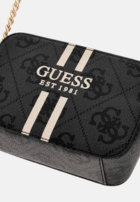 Black patterned handbag with gold chain and prominent white stripes, featuring the "GUESS EST 1981" logo. Textured material, rectangular shape.