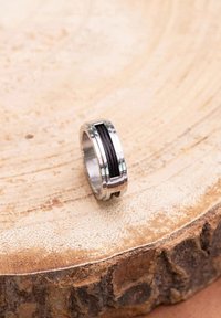Silver ring featuring a central black textured band, with polished edges and grooves. Displayed on a wooden surface.