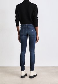 Jeans ajustados de color azul con cintura alta, que presentan un sutil logo en el bolsillo trasero, combinados con botas blancas hasta el tobillo y un suéter negro.