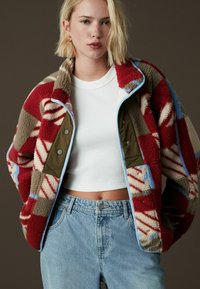Veste en polaire rouge et grise avec des motifs abstraits, bord bleu et boutons-pression, portée sur un top court blanc et un jean bleu clair.
