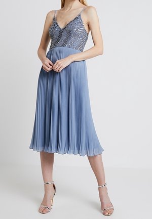Ærmeløs sølvfarvet paillet-top med plisseret lyseblåt midinederdel, båret med sølvfarvede åbne højhælede sandaler.