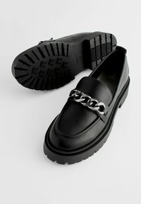 Zwarte leren loafers met een stevige rubberen zool en een decoratieve zilveren ketting over de bovenkant. Soepele textuur met een afgerond neusstuk.