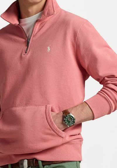 Jasnoróżowa bluza z zamkiem na szyi, z kieszenią typu kangur i ściągaczami na rękawach. Posiada małe haftowane logo na klatce piersiowej. Gładka tekstura materiału.