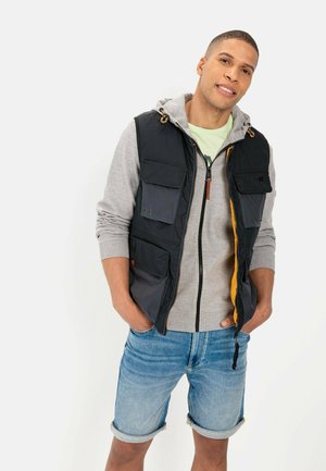 Marineblå vest med flere lommer over en grå hættejakke, parret med opkrøllede denimshorts. Har lynlås lukning og lysebrune detaljer.