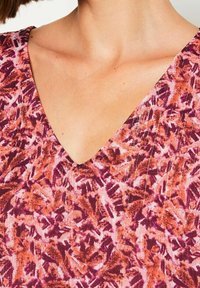 V-pääntie yläosa, jossa on elinvoimainen pinkki ja oranssi abstrakti kukkakuosi ja teksturoitu pinta, joka korostaa herkkiä kankaan yksityiskohtia.