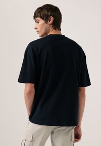 Młody mężczyzna z krótkimi ciemnymi włosami, ubrany w gładki czarny t-shirt i jasnobeżowe spodnie typu cargo, zwrócony tyłem i patrzący na bok.