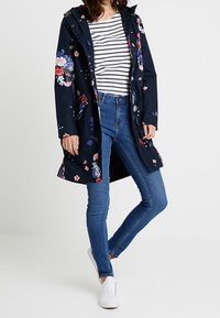 Marineblomstret regnfrakke med hætte, prydet med blomstermotiv, parret med en sort og hvid stribet top og blå skinny jeans. Hvite sneakers.