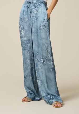 Frau trägt lockere, weit geschnittene blaue Hose mit Blumenmuster, elastischem Bund mit Kordelzug und gewebten Sandalen mit offenen Zehen, steht mit Händen in den Taschen.