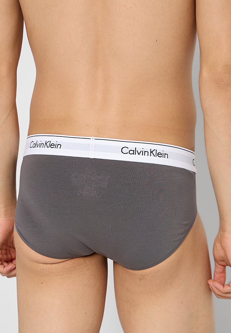 Calzoncillos de algodón gris con una cinturilla blanca que presenta la marca "Calvin Klein" en negro. Textura suave, diseño ajustado y corte bajo.