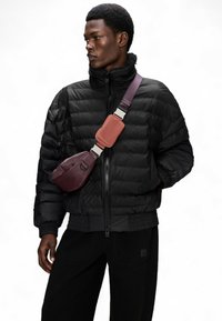 Schwarze Pufferjacke mit hohem Kragen, kombiniert mit einer Gürteltasche in einem burgunderroten und rosa Farbschema. Strukturiertes, dickeres Gewebe.