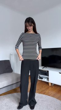 Μπλούζα με μακρύ μανίκι σε ασπρόμαυρες ριγέ, συνδυασμένη με άνετο μαύρο παντελόνι, φορεμένη με μαύρα παπούτσια, πάνω σε φωτεινό εσωτερικό φόντο.