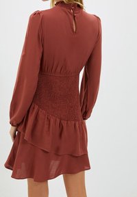 Rostfarbendes Kleid mit gerafftem Bund, langen Ärmeln und schichteter Rock. Verfügt über einen Schlüsselanhauschnitt am Rücken mit Knopfdetails. Glatte Stoffstruktur.