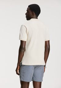 Világos bézs színű pólóing, texturált anyagból, rövid ujjú, hajtott gallérral, navy és fehér csíkos rövidnadrággal párosítva.