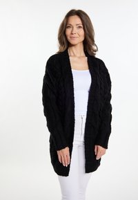 Cardigan nero lavorato a maglia con motivo a treccia, apertura frontale, maniche lunghe e vestibilità rilassata, indossato sopra una maglietta bianca e pantaloni chiari.