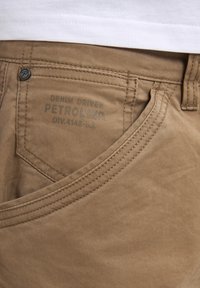Pantalones de mezclilla beige con una textura suave. Presenta un bolsillo estructurado y un botón de metal. Logo "Petrol Industries" grabado en la cinturilla.