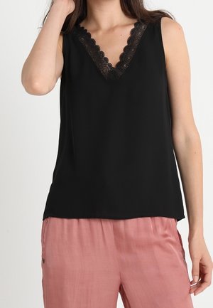 Vrouw draagt een zwart mouwloos topje met kanten V-hals en losse roze broek, poseert met één hand haar haar aanrakend.