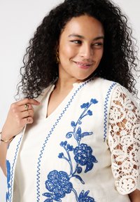 Witte top met blauwe bloemenborduursels, gekartelde randen en kantdetails op de mouwen. Heeft een V-hals en een gestructureerde stof.