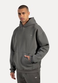 Grauer Hoodie aus weichem Material, mit einer Fronttasche, gerippten Bündchen und einem kleinen Logopatch auf der Brust. Schlichtes, entspanntes Design.