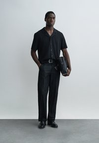 Homme portant une chemise noire à manches courtes et des pantalons noirs texturés, tenant un sac noir, debout devant un fond gris uni.