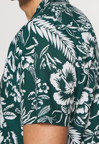 Lyhythihainen t-paita tummanvihreästä kankaasta, jossa on valkoisia kukka- ja lehtikuoseja. Sujuva pinta ja rento leikkaus, näkyvä kaulus ja napit.