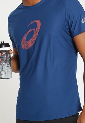 Mand iført en blå Asics t-shirt, der holder en klar vandflaske med hvidt og sort låg på en hvid baggrund.
