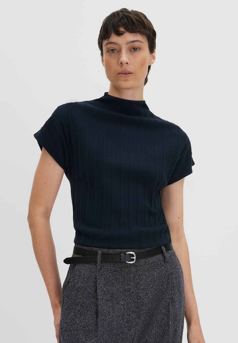 Top en tricot côtelé bleu marine avec des manches courtes roulées et un col montant, associé à un pantalon gris texturé et une ceinture noire en accent.