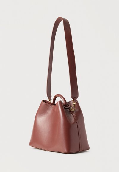 Καφέ δερμάτινη τσάντα τύπου κάδο με μακρύ λουρί ώμου, μικρή λαβή και χρυσά μεταλλικά στοιχεία, που στέκεται σε λευκό φόντο.