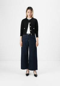 Femme se tenant contre un fond blanc, portant un cardigan noir court avec des boutons blancs, un haut blanc, un jean en denim foncé à jambes larges, et des talons noirs.
