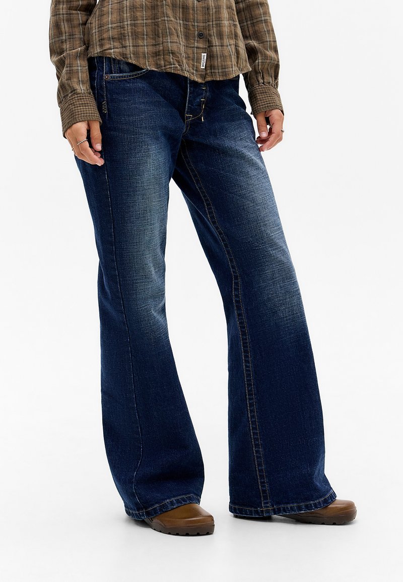 Donkerblauwe denim wijduitlopende jeans met subtiele vervaging, een middelhoge taille en contrasterende stiksels. Gepaard met een bruine geruite shirt en bruine schoenen.