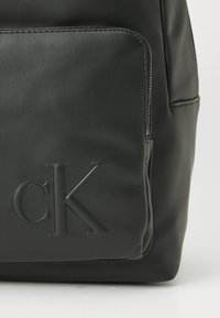 Mochila de cuero negro con un bolsillo que tiene el logo "cK" embellecido en una superficie lisa y texturizada, y con una cremallera visible sobre fondo blanco.
