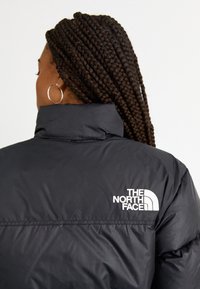 Personne aux cheveux tressés portant une doudoune noire The North Face, vue de dos sur un fond uni clair.