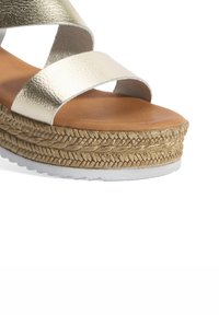 Sandale à lanières métalliques dorées avec une semelle intérieure en cuir marron, un intermédiaire en jute tressé et une semelle extérieure en caoutchouc blanc, présentant un design à talon compensé.