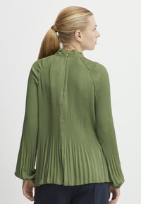 Blusa verde plissettata con colletto alto, maniche lunghe e chiusura con bottoni sul retro. Presenta un tessuto testurizzato e leggero.