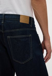 Jeans de mezclilla de color azul oscuro con un parche de cuero marrón claro en la cintura etiquetado como "WEEKDAY". Presenta costuras dobles en los bolsillos y una textura suave.