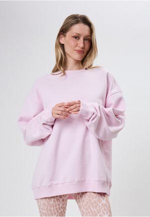 Kobieta z blond włosami do ramion, ubrana w oversize jasnoróżową bluzę i beżowe legginsy w panterkę, stojąca na jednolitym tle.