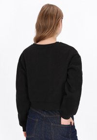 Fekete, rövidített pulóver hosszú ujjakkal, kerek nyakkivágással és puha textúrával. Sötét farmernadrággal viselve, hátul részletek láthatók.