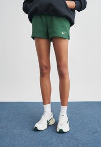 Πράσινα βαμβακερά σορτς με λευκό λογότυπο Nike· συνδυασμένα με λευκές κάλτσες και παπούτσια με πράσινες λεπτομέρειες σε μπλε πάτωμα.