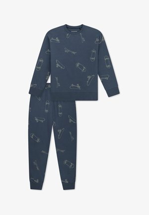Bambino sorridente in piedi a piedi nudi che indossa un pigiama blu scuro abbinato con leggeri motivi di skateboard su maglia e pantaloni.