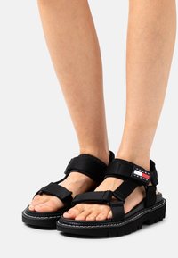 Svarta remsandaler med en kraftig sula, utrustade med justerbara kardborreband. Anmärkningsvärd röd och vit logotyp som accent på en av remmarna.