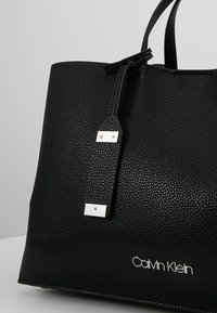 Czarna skórzana torba typu tote z teksturowaną powierzchnią, z metalowymi elementami w srebrnym kolorze oraz logo. Design obejmuje mocne uchwyty i ustrukturyzowany kształt.